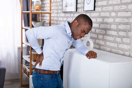 Side View Of A Young Man Leaning On Cabinet Suffering From Back Painの写真素材