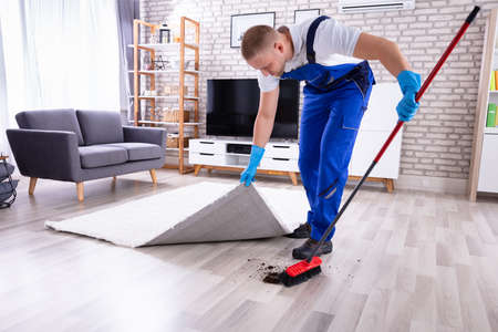 Young Male Janitor In Uniform Removing Dirt Under The Carpetの写真素材