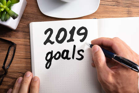 Close-up Of A Person's Hand Writing 2019 Year Goals On Dotted Notebook Over Wooden Desk