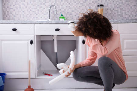 Close-up Of A Young African Woman Looking At Broken White Sink Pipe In The Kitchenの写真素材
