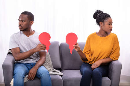 Couple Sitting On Sofa Ignoring Each Other And Holding Broken Red Heart