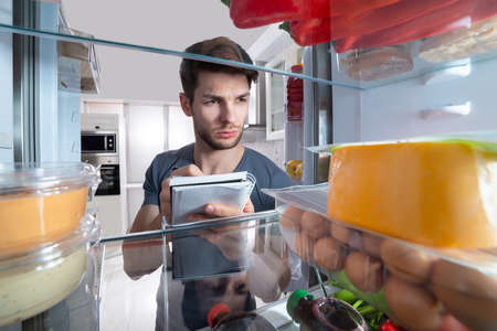 Close-up Of Man Writing On Spiral Book Near Open Refrigerator In Kitchen