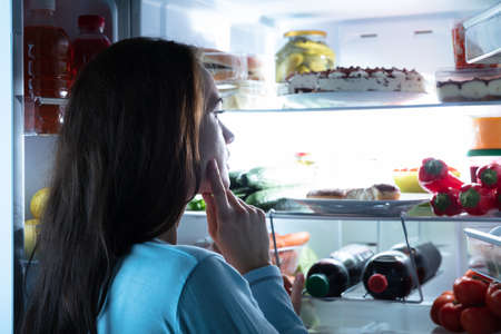 Close-up Of A Thoughtful Young Woman Looking For Food In An Open Refrigeratorの写真素材