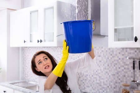 Unhappy Young Woman Collecting Water Leaking From Ceiling In Blue Bucket