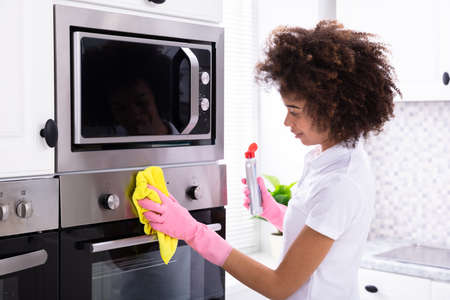 Close-up Of A Smiling Female Janitor Cleaning Oven With Yellow Napkin In The Kitchenの写真素材