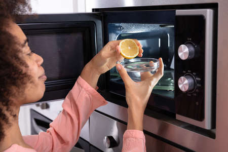 Woman's Hand Squeezing Halved Lemon In The Glass Bowl Near An Open Microwave Ovenの写真素材