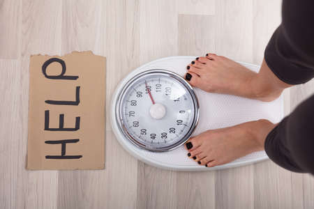 Elevated View Of Help Sign On Cardboard With Woman's Feet On Weighing Scale