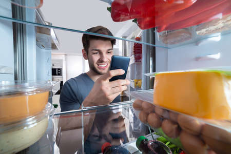 Happy Man Standing In Front Of Refrigerator Using Mobilephone In Kitchenの写真素材