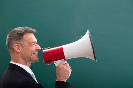Happy Mature  Man Doing Announcement Using Megaphone In Front Of Chalkboardの写真素材