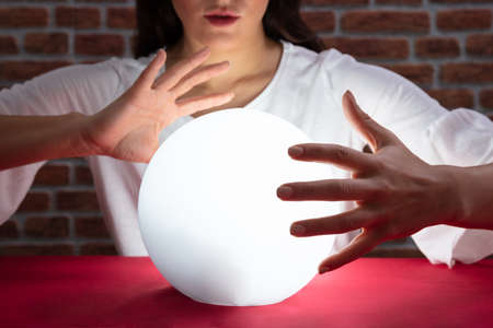 Close-up Of Fortuneteller's Hand Covering The Glowing Crystal Ball On Red Deskの写真素材