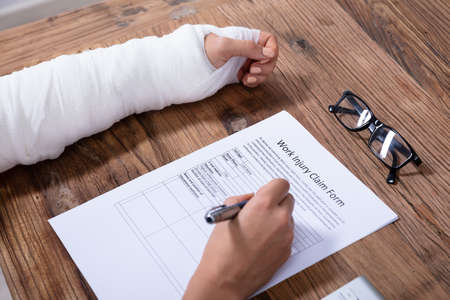Close-up Of Businesswoman With White Bandage Hand Filling Work Injury Claim Form On Wooden Deskの写真素材