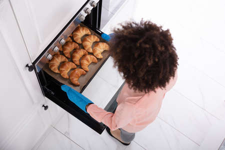 A Happy Young Woman Wearing Blue Mitt Removing Baked Croissants Tray From An Oven