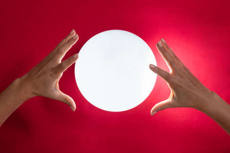 Close-up Of Fortuneteller's Hand Covering The Glowing Crystal Ball On Red Desk