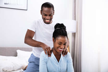 Happy Young African Man Giving Shoulder Massage To His Wife In Bedroomの写真素材