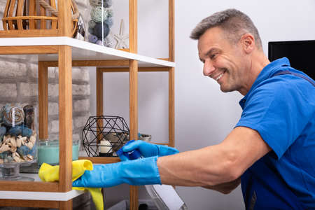 Smiling Mature Male Janitor Cleaning Shelf With Napkin And Detergent Spray Bottle