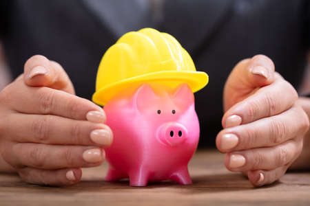 Close-up Of A Person's Hand Protecting Pink Piggy Bank With Yellow Hard Hatの写真素材