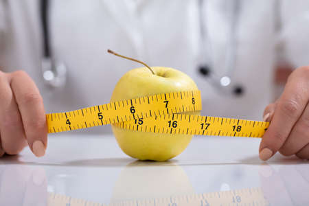 Close-up Of Female Dietitian Measuring The Whole Apple With Measuring Tape On Deskの写真素材