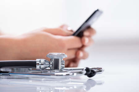 Close-up Of Stethoscope In Front Of Woman's Hand Holding Cellphone Over Reflective White Deskの写真素材