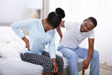 African Couple Suffering From Back Pain Sitting On Bed