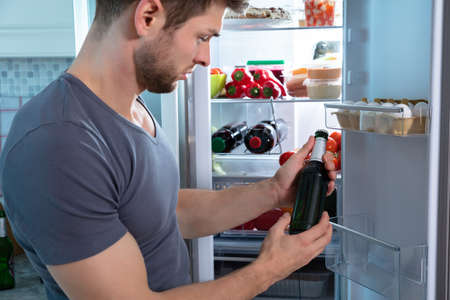 Young Man Holding Beer Bottle Standing In Front Of Refrigeratorの写真素材