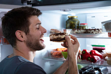 Portrait Of A Happy Man Eating Cake Near Open Refrigeratorの写真素材