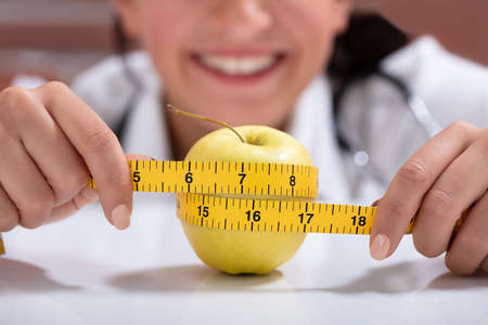 Close-up Of Female Dietitian Measuring The Whole Apple With Measuring Tape On Deskの写真素材
