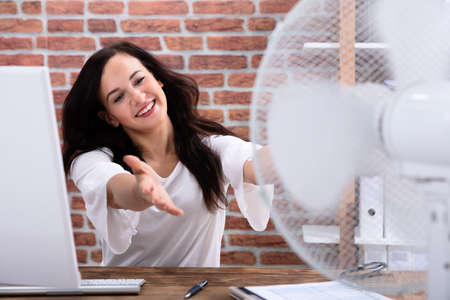 Happy Mature Businesswoman Cooling Herself With Electrical Fan At Workplace On Wooden Deskの写真素材