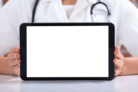 Close-up Of Female Doctor's Hand Holding Digital Tablet With Blank White Screen On White Deskの写真素材