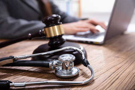 Close-up Of Mallet And Stethoscope On Gavel Over The Office Desk With Businessperson Using Laptopの写真素材