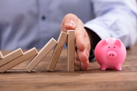 Close-up Of A Businessperson's Hand Protecting House Model From Falling Wooden Blocks On Desk