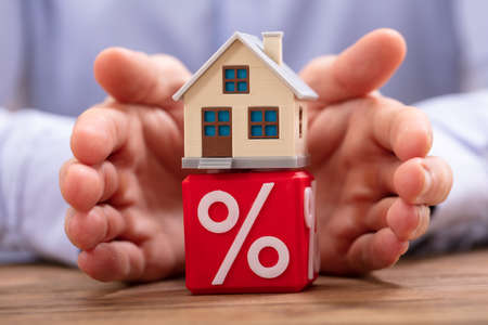 Close-up Of A Person's Hand Protecting House Model Over Red Cube With Percentage Symbolの写真素材