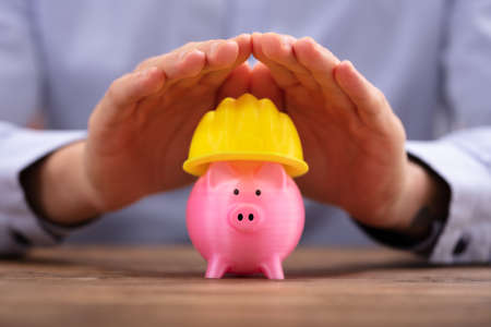 Close-up Of A Person's Hand Protecting Pink Piggy Bank With Yellow Hard Hatの写真素材