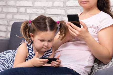 Close-up Of Mother And Daughter Sitting On Sofa And Using Mobilephone At Homeの写真素材