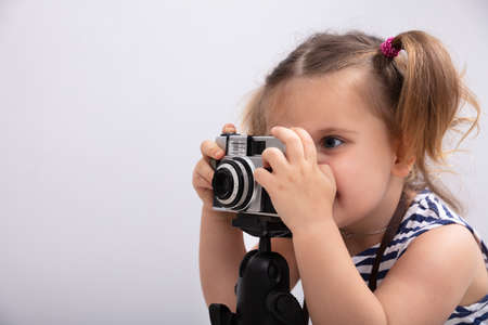 Little Girl Standing Against White Background While Taking Picture Using Photo Cameraの写真素材
