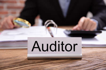 Nameplate With Auditor Title Kept On Desk In Front Of Businessman Examining The Invoicesの写真素材