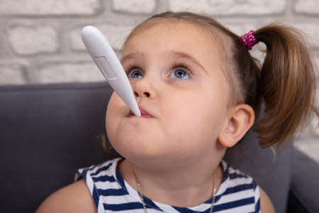 Sick Girl Sitting On Sofa With A Thermometer In Her Mouth Measuring The Feverの写真素材