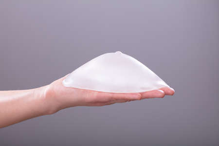 Close-up Of Woman's Hand Holding Silicon Breast Implant Against Gray Background