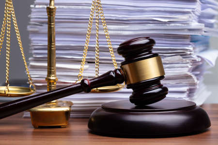 Justice Scales And Wooden Gavel In Front Of Stacked Paper On Wooden Desk