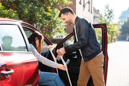 Smiling Young Husband Helping Her Disabled Wife With Crutches To Get Inside The Carの写真素材