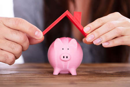 Human Hand Protecting Pink Piggybank With Red Roof Over Wooden Deskの写真素材