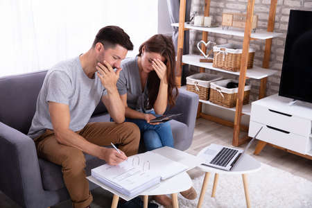 Young Sad Couple Sitting On Sofa Calculating Invoice