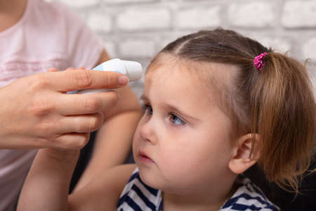 Mother Checking Fever Of Her Sick Daughter Using Forehead Thermometer At Home