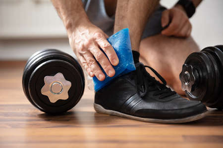 Low Section Of A Man Applying Ice Gel Pack On An Injured Ankle In Gymの写真素材
