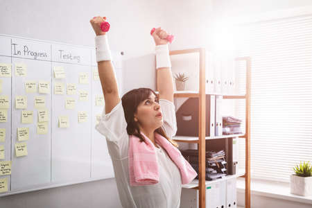 Smiling Young Businesswoman Exercising With Red Dumbbells In Office