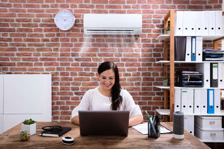 Smiling Businesswoman Enjoying The Cooling Of Air Conditioner Using Laptop At Workplace