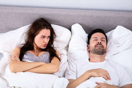 Angry Young Woman Looking At Man Snoring In Bed At Home