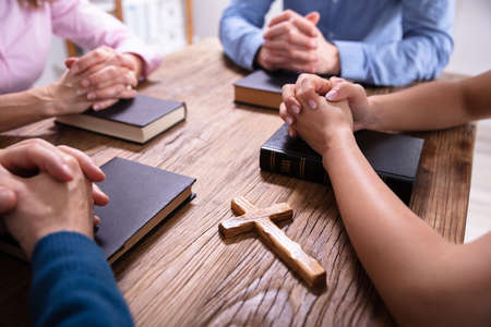 Close-up Of Businesspeople's Praying Hands Over The Bible On Wooden Deskの写真素材