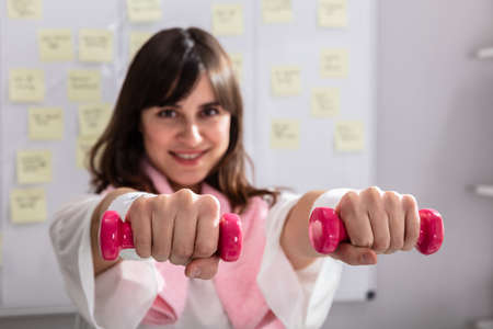 Smiling Young Businesswoman Exercising With Red Dumbbells In Office