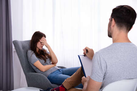 Male Psychologist With Clipboard Sitting Near Young Woman Suffering From Depressionの写真素材