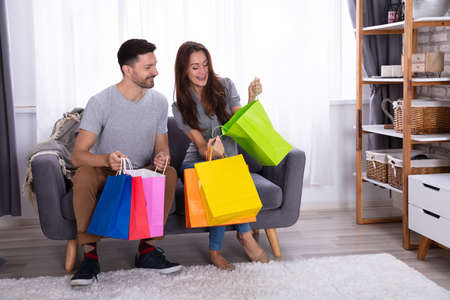 Happy Couple Sitting On Sofa Looking In Shopping Bag At Homeの写真素材